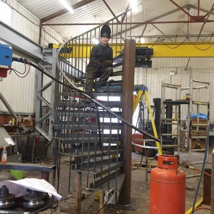 Spiral staircase being built in a workshop, with a welder standing on the steps